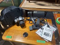 Canon EOS Rebel 300D camera body with attached Canon Zoom Lens EF-S 18-55mm 1:3.5-5.6 and included accessories on wooden table. Bag, charger, batteries, manuals, and lens cap visible.
