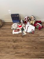 Wide view of all Christmas items including two Santa figurines, blue advent calendar, folded Christmas towels and tea towels with festive prints, red Christmas sweater, and other holiday decor items arranged on wooden floor.