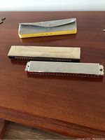 Two harmonicas alongside a gray case on a wooden surface. One harmonica is silver colored with some rust, the other has a red comb. Gray case is fold-over style with Hohner gold lettering.