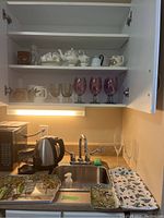 Photo showing kitchen cupboard open with seated shelves displaying Foley China white porcelain tea set including teapot, creamer, sugar bowl, saucers and teacups; Bunnykins mug; multi-colored glass and vintage style purple goblets; melamine trays with floral prints on counter below.
