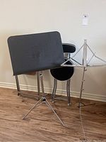 Photo showing one black padded folding chair behind a black perforated sheet music holder on adjustable metal tripod stand.