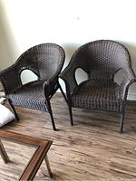 Two dark brown wicker patio chairs side by side on a wood floor against a white wall.