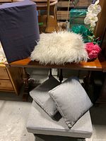 Light gray ottoman with two matching throw pillows, white shaggy throw pillow, blue wedge pillow, and fake flowers on wood table