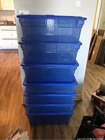 Stack of seven blue Rubbermaid storage containers with lids. Containers are translucent blue plastic and stacked near a wall.