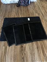 Photo showing four small black rubber backed rugs placed on a larger blue or dark throw rug on hardwood floor.