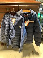 Two men's winter jackets hanging on wooden hangers in front of a wooden dresser, showing overall look and details such as tags and reflective strips.