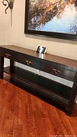 Full view of the wooden console table showing three drawers and a bottom shelf, placed on a wooden floor under a framed artwork.