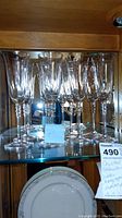 Photo showing a set of 8 clear crystal stemware glasses displayed on a glass shelf, including taller water glasses and slightly shorter champagne glasses with visible decorative stem detail.