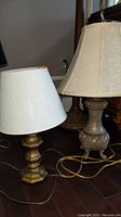 Two metal table lamps with cream fabric shades on floor, showing relative sizes and bases (one geometric brass base, one ornate bronze-toned base)