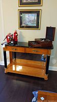 View of wooden server with black marble top, two drawers with brass handles, black legs accented with gold, lower shelf, and mirrored back, displaying figurines and a wooden box on top.