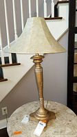 Full view of the table lamp standing on a round table, showing the faux gold metal finish on the tall base and the flared beige lampshade.