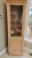 Front view of tall wooden cabinet with glass door showing three glass shelves inside and wooden lower door.