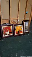 Three framed wall art pieces leaning against an unfinished wall; two square floral paintings with dark wood frames and one rectangular print with gold-toned frame and linen matting.