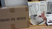 Photo of cardboard box labeled Smash Ice Device and Oster 8-speed blender box beside a pre-owned Oster blender unit.