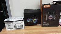 Photo showing boxed items: one white Ascent CD Boombox, one black boombox, and Carise T26 Pro karaoke speaker box