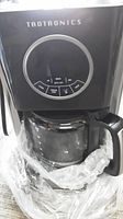 Close-up image of black Taotronics coffee maker with glass carafe inside a plastic bag, showing control dial with brew settings.