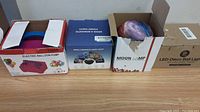 Four boxed items including an electric balloon pump, a bubble machine, a moon lamp, and a LED disco ball light on a floor against a beige wall.