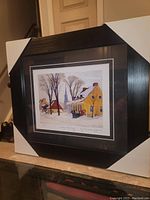 View of full framed and matted print in black frame with white matting, showing winter village scene.