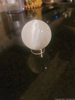White polised selenite sphere on a clear plastic stand shown on a dark surface
