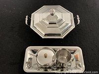 Octagonal stainless steel serving dish with a lid featuring a decorative knob and two side handles on a black surface.