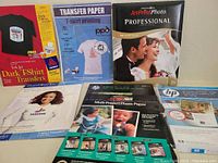 Photo showing six boxes of various iron-on transfer papers and glossy photo paper, including branded boxes from JetPrint, HP, POD, ARK, and Voodoo Graphics for T-shirt printing and photo printing.