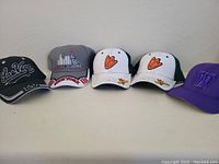Five baseball-style caps arranged in a row. From left to right: black Las Vegas hat, gray New York City hat, two white and green University of Oregon Ducks hats with orange duck footprints, and purple University of Washington hat.