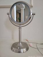 Front view of the two-sided makeup mirror showing the normal side, mounted on a silver-tone pedestal stand with a round base and a button.