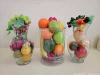 Photo of three decorative glass jars each filled with various life-size artificial fruits including grapes, apples, pears, oranges, bananas, and mixed fruits, arranged side by side on a flat surface.