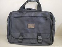 Front view of a black travel bag with handle and front flap secured by two black quick-release buckles. A patch with letters is centered on the front flap.