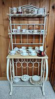 Full view of the vintage baker's rack showing wooden prep surface with various teacups and dishes on shelves, white metal frame with scroll detail.