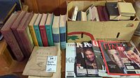 Overview photo showing a variety of books and magazines placed on a table and inside a partly open cardboard box. Books include several with red spines, colorful hardcovers, and magazines with JFK Jr on cover.
