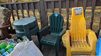 View of the dark green Adirondack chairs, round plastic side tables, and white high back patio chairs stacked.