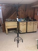 Black metal coat rack with hooks and umbrella ring standing in a basement with wood paneling and vintage cabinets in the background.