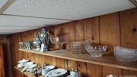 Wide view showing multiple items including metal cacao pot, teapot, glass stemware, and dishware arranged on wooden shelf