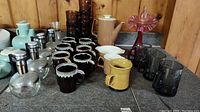 Photo showing assortment of cups, carafe, mid-century pitcher set, and cranberry vase on table with wood panel background