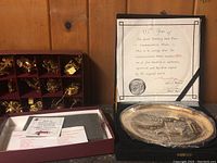 Photo of commemorative plate wrapped in plastic alongside the certificate and a box containing multiple Christmas tree ornaments