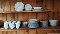 Full shelf view showing stacks of plates, teacups, and saucers on a wood shelf against wood paneling.