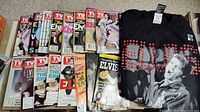 Photo showing 13 Elvis Presley TV Guide magazines laid out in two rows with the XL Elvis t-shirt folded beside them. Magazines have various Elvis-themed covers including one with a hologram and multiple mini CDs attached.