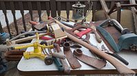 Overview of various weathered garden tools on a white tabletop showing sprinklers, clippers, trowels, ax, saws and bug zapper