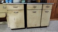 Front view of three vintage Geneva metal kitchen cabinets with cream finish showing rust and wear. Each cabinet has metal handles on drawer and cupboard.