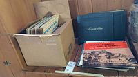Box filled with multiple vinyl LP records and some albums stacked beside it including a record album book