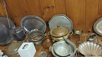 Overview of mixed metalware items including hammered aluminum roasting pots and Dutch ovens with glass lids, ceramic tissue box cover, and round trays.