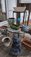 Multiple planters with various shapes including ceramic cream-colored rectangular planter and green oval planter on black metal plant stands, with garden figurines in front.