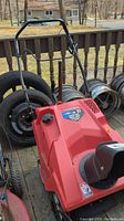 Front view of the red Troy-Bilt 21-inch snow blower with black handle, snow discharge chute facing forward, surrounded by tires and rims on wooden deck.