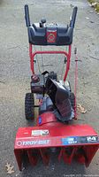 Front view of the Troy-Bilt Storm 5024 snow blower showing red metal body, intake augers, and control handles.