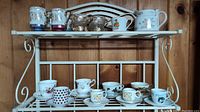 White metal rack with two shelves displaying assorted teacups and saucers. Top shelf has silver and decorated blue/white mugs and small creamers. Bottom shelf shows various teacups with patterns including hearts and U.S. state maps.