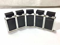 Five clear glass rectangular perfume bottles with black opaque fronts and black square caps arranged in a row on a white fabric surface, front view showing the printed text.