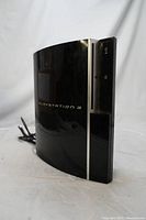 Oblique angled front view of glossy black PS3 console showing front, disc drive, and power cable extending from back.