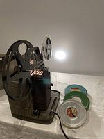 Image of Bell & Howell portable movie projector with two large reels and power cord, alongside three metal film reels in silver and green with vintage labels.
