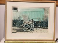 Framed serigraph artwork showing a rocking chair covered with a crocheted blanket and a cat sitting on it, in a garden setting with a mint green wooden fence and flowers.